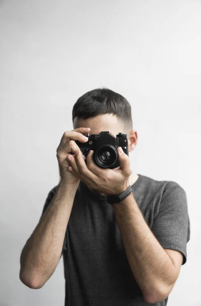 Beau jeune homme barbu dans une chemise noire tenant vintage caméra de film à l'ancienne sur un fond blanc et regardant dans le viseur de la caméra . — Photo