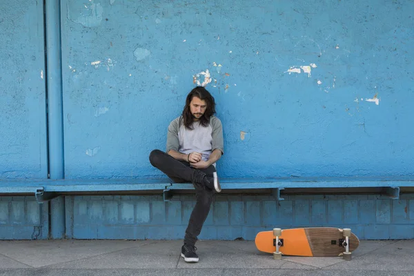 Skater sitzt auf blauem Hintergrund mit orangefarbenem Skateboard in seiner Nähe. — Stockfoto