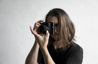 Yakışıklı genç sakallı adam uzun saçlı ve vintage eski moda film kamera beyaz bir arka plan üzerinde tutarak ve vizör kamera seyir siyah gömlekli.