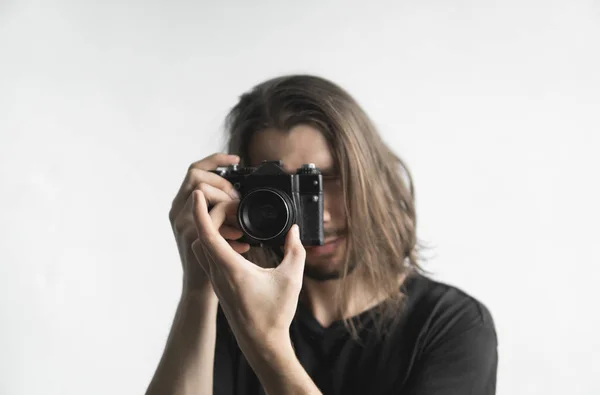 Bonito jovem barbudo homem com um cabelo longo e em uma camisa preta segurando câmera de filme antiquado vintage em um fundo branco e olhando no visor da câmera . — Fotografia de Stock