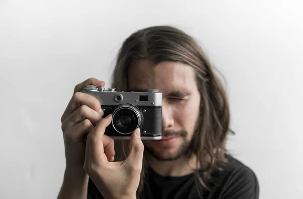 Bonito jovem barbudo homem com um cabelo longo e em uma camisa preta segurando câmera de filme antiquado vintage em um fundo branco e olhando no visor da câmera . — Fotografia de Stock