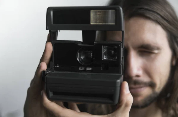 Pohledný mladý vousatý muž s dlouhými vlasy a v černé košili drží fotoaparát vintage staromódní film na bílém pozadí a při pohledu do fotoaparát hledáček. — Stock fotografie