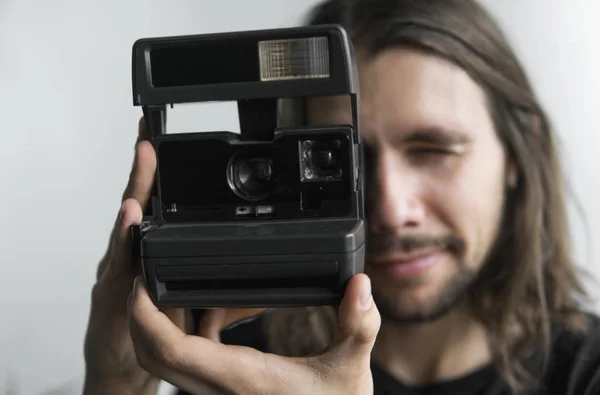 Bonito jovem barbudo homem com um cabelo longo e em uma camisa preta segurando câmera de filme antiquado vintage em um fundo branco e olhando no visor da câmera . — Fotografia de Stock