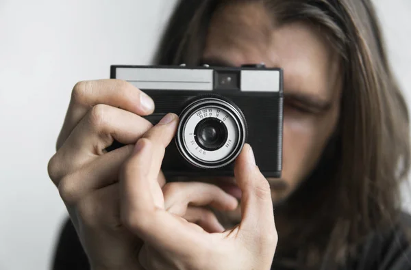 Bonito jovem barbudo homem com um cabelo longo e em uma camisa preta segurando câmera de filme antiquado vintage em um fundo branco e olhando no visor da câmera . — Fotografia de Stock