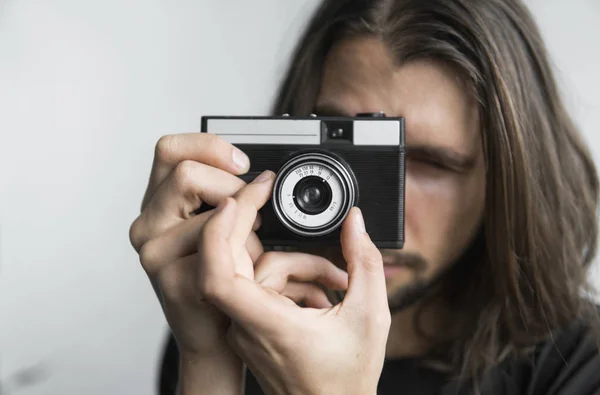 Bonito jovem barbudo homem com um cabelo longo e em uma camisa preta segurando câmera de filme antiquado vintage em um fundo branco e olhando no visor da câmera . — Fotografia de Stock