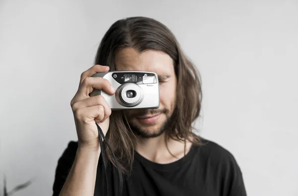 Bonito jovem barbudo homem com um cabelo longo e em uma camisa preta segurando câmera de filme antiquado vintage em um fundo branco e olhando no visor da câmera . — Fotografia de Stock