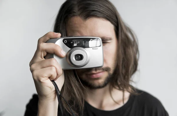 Bonito jovem barbudo homem com um cabelo longo e em uma camisa preta segurando câmera de filme antiquado vintage em um fundo branco e olhando no visor da câmera . — Fotografia de Stock