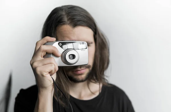 Bonito jovem barbudo homem com um cabelo longo e em uma camisa preta segurando câmera de filme antiquado vintage em um fundo branco e olhando no visor da câmera . — Fotografia de Stock