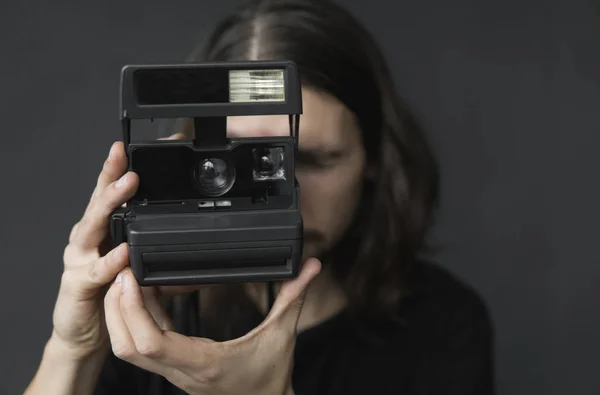 Yakışıklı genç sakallı adam uzun saçlı ve vintage eski moda film kamera siyah bir arka plan üzerinde tutarak ve vizör kamera seyir siyah gömlekli. — Stok fotoğraf