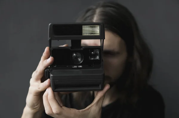 Bonito jovem barbudo homem com um cabelo longo e em uma camisa preta segurando câmera de filme antiquado vintage em um fundo preto e olhando no visor da câmera . — Fotografia de Stock