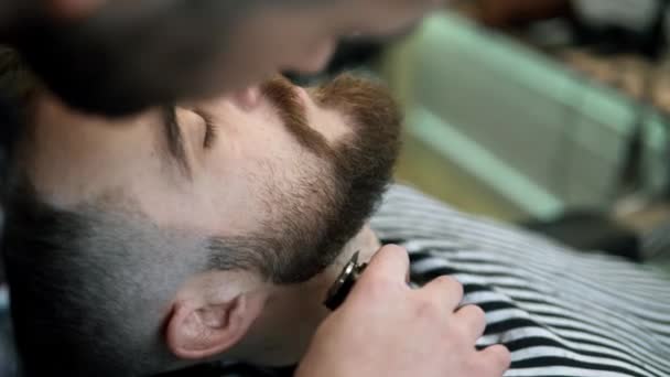 Ухаживаю за бородой. Парикмахерская. Мужчина парикмахер делает стрижку бороды взрослых мужчин в мужском парикмахерской с электрическим клиппером. Красивый молодой человек стрижется в парикмахерской . — стоковое видео