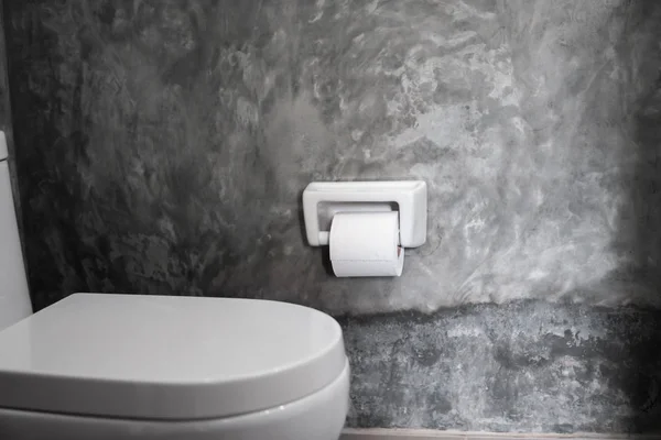 Белое подвешенное сиденье унитаза на белом унитазе в домашней ванной комнате с серой плиткой в бетонном стиле и туалетной бумагой на стене. Ванная комната люкс . — стоковое фото