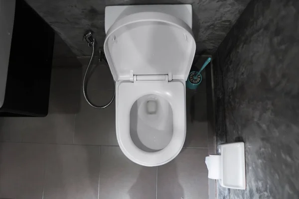 Λευκό κρέμεται κάθισμα τουαλέτας σε λευκή τουαλέτα στο σπίτι μπάνιο με γκρι πλακάκια σε σκυρόδεμα και χαρτί τουαλέτας στον τοίχο. Μπάνιο πολυτελείας εσωτερικό. — Φωτογραφία Αρχείου