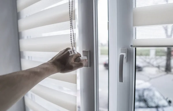 Man holding window handle on a plastic window with white fabric roller blinds in the living room. Close up on roll curtains indoor.