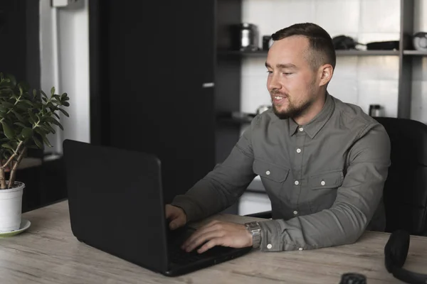 Ευτυχισμένο χαμόγελο νέος επιχειρηματίας ή φοιτητής σε ένα πουκάμισο που κάθεται ενάντια στην οθόνη του υπολογιστή. Εργασία σε έναν υπολογιστή σε έναν πίνακα στο γραφείο με μια προσεκτική έκφραση. — Φωτογραφία Αρχείου