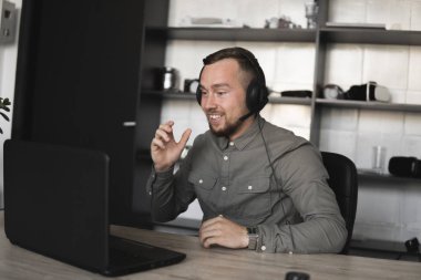 Mutlu gülümseyen Genç işadamı ya da öğrenci bir gömlek kulaklık bilgisayar monitörü karşı oturan ve internet üzerinden biriyle konuşan. Ofisteki bir masada bir bilgisayar üzerinde çalışmak.