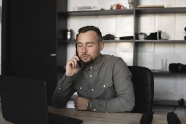 Genç işadamı veya öğrenci bir gömlek bilgisayar monitörü karşı oturan ve akıllı telefon ile iş ortağı ile konuşuyor. Düşünceli bir ifade ile ofiste bir masada bir pc üzerinde çalışma.