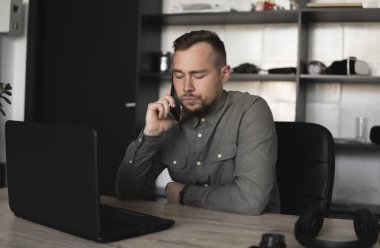 Genç işadamı veya öğrenci bir gömlek bilgisayar monitörü karşı oturan ve akıllı telefon ile iş ortağı ile konuşuyor. Düşünceli bir ifade ile ofiste bir masada bir pc üzerinde çalışma.