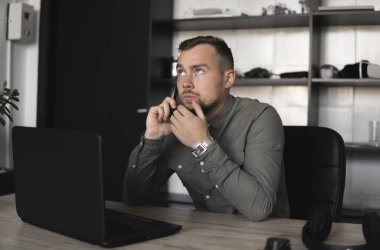 Genç işadamı veya öğrenci bir gömlek bilgisayar monitörü karşı oturan ve akıllı telefon ile iş ortağı ile konuşuyor. Düşünceli bir ifade ile ofiste bir masada bir pc üzerinde çalışma.