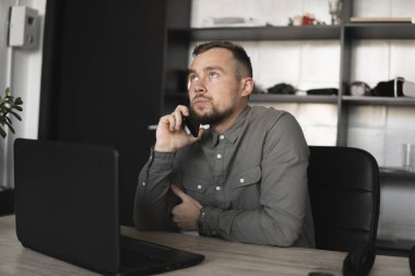 Genç işadamı veya öğrenci bir gömlek bilgisayar monitörü karşı oturan ve akıllı telefon ile iş ortağı ile konuşuyor. Düşünceli bir ifade ile ofiste bir masada bir pc üzerinde çalışma.
