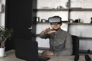Sanal gerçeklik gözlükleriyle bilgisayara karşı oturan genç sakallı adam bir şeye dokunmaya ya da gördüklerinin önünde bir şeye dokunmaya çalışıyor. Modern teknolojiler. Geleceğin teknolojisi kavramı.