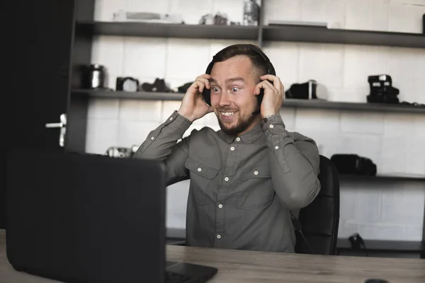 Εντυπωσιασμένος χαρούμενος χαμογελαστός νέος επιχειρηματίας ή φοιτητής σε ένα πουκάμισο που κάθονται ενάντια στην οθόνη του υπολογιστή σε ακουστικά και να μιλήσει με κάποιον μέσω του Διαδικτύου. Εργασία σε έναν υπολογιστή σε ένα τραπέζι στο γραφείο. — Φωτογραφία Αρχείου