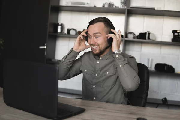 Εντυπωσιασμένος χαρούμενος χαμογελαστός νέος επιχειρηματίας ή φοιτητής σε ένα πουκάμισο που κάθονται ενάντια στην οθόνη του υπολογιστή σε ακουστικά και να μιλήσει με κάποιον μέσω του Διαδικτύου. Εργασία σε έναν υπολογιστή σε ένα τραπέζι στο γραφείο. — Φωτογραφία Αρχείου