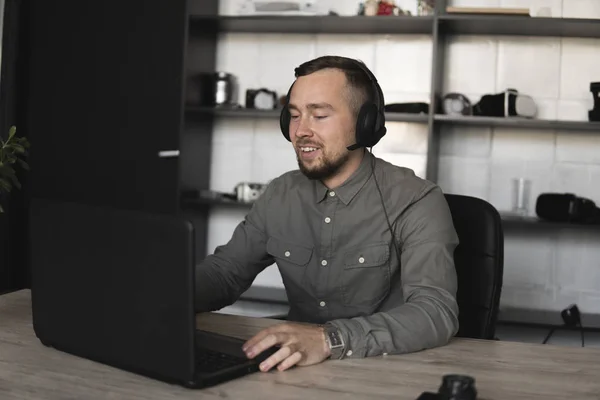 Νέος επιχειρηματίας ή μαθητής σε ένα πουκάμισο που κάθεται ενάντια στην οθόνη του υπολογιστή στα ακουστικά. Εργασία σε έναν υπολογιστή σε έναν πίνακα στο γραφείο με μια προσεκτική έκφραση. — Φωτογραφία Αρχείου