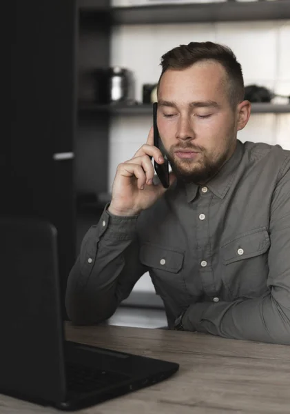 Νέος επιχειρηματίας ή μαθητής σε ένα πουκάμισο που κάθεται ενάντια στην παρακολούθηση του υπολογιστή και μιλά με τον συνεργάτη του από το smartphone. Εργασία σε έναν υπολογιστή σε έναν πίνακα στο γραφείο με μια προσεκτική έκφραση. — Φωτογραφία Αρχείου