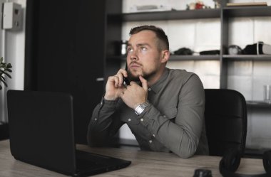 Genç işadamı veya öğrenci bir gömlek bilgisayar monitörü karşı oturan ve akıllı telefon ile iş ortağı ile konuşuyor. Düşünceli bir ifade ile ofiste bir masada bir pc üzerinde çalışma.