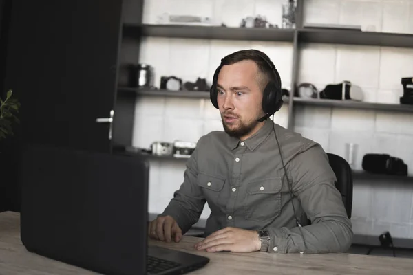 Μπερδεμένος ή θυμωμένος επιχειρηματίας ή μαθητής σε ένα πουκάμισο που κάθεται ενάντια στην οθόνη του υπολογιστή στα ακουστικά και μιλώντας με κάποιον μέσω Διαδικτύου. Εργασία σε έναν υπολογιστή σε ένα τραπέζι στο γραφείο. — Φωτογραφία Αρχείου