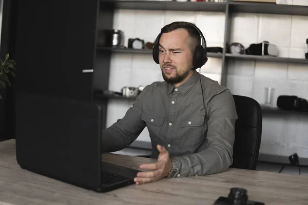Confused or angry businessman or student in a shirt sitting against monitor of computer in headphones and speaking with somebody via internet. Working on a pc at a table in the office.