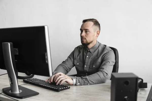 Νέος επιχειρηματίας ή μαθητής σε ένα πουκάμισο που κάθεται ενάντια στην οθόνη του υπολογιστή. Δουλεύοντας σε έναν υπολογιστή σε ένα τραπέζι στο γραφείο με μια προσεκτική έκφραση. Νέος επιχειρηματίας που εργάζεται για το laptop του στο γραφείο. — Φωτογραφία Αρχείου
