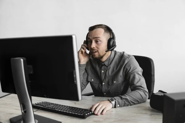 Νέος επιχειρηματίας ή μαθητής σε ένα πουκάμισο που κάθεται ενάντια στην οθόνη του υπολογιστή με ακουστικά και μιλά με κάποιον μέσω Διαδικτύου. Εργασία σε έναν υπολογιστή σε ένα τραπέζι στο γραφείο. — Φωτογραφία Αρχείου