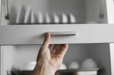 El, kurutma makinesinin kapısını çatal bıçakla tutuyor. Tabak kurutucu metal raf ve beyaz temiz tabaklar. Geleneksel rahat mutfak. Islak tabak ve kâselerle dolu beyaz tabak boşaltma dolabını aç..