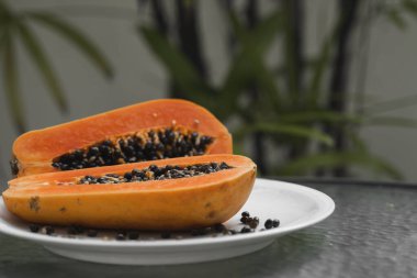 Beyaz tabakta tohum ve arka planda yeşil bitkiler olan olgun papayaların yarısı. Tatlı papaya dilimleri. Yarım papayalar. Sağlıklı egzotik meyveler. Vejetaryen yemeği..
