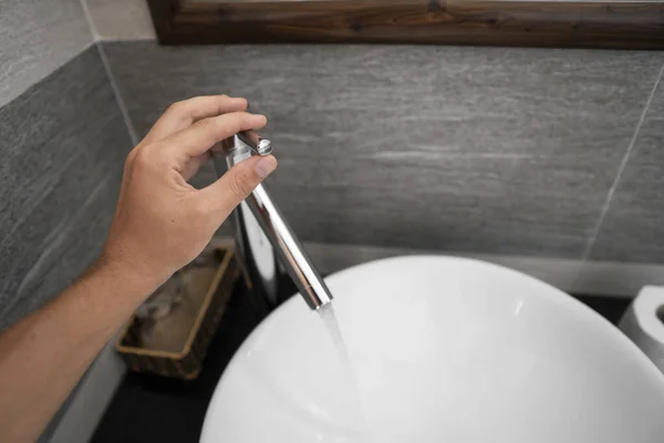 La mano masculina usa un grifo en el interior de un baño con lavabo redondo blanco y grifo de cromo. Agua que fluye del grifo de cromo . — Foto de Stock