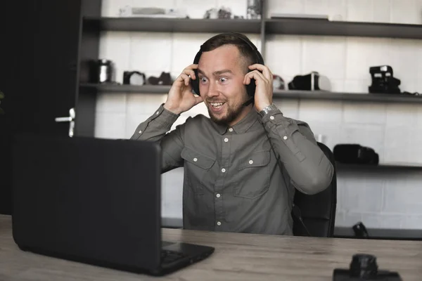 Χαρούμενος χαμογελαστός νέος επιχειρηματίας ή φοιτητής σε ένα πουκάμισο που κάθεται ενάντια στην οθόνη του υπολογιστή με ακουστικά και μιλώντας με κάποιον μέσω του Διαδικτύου. Εργασία σε έναν υπολογιστή σε ένα τραπέζι στο γραφείο. — Φωτογραφία Αρχείου