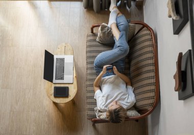 Bir kadın kanepede dizüstü bilgisayar ve masada kitap okurken akıllı telefon kullanır. Çevrimiçi çalışma, serbest çalışma. Serbest çalışan ya da serbest çalışan bir kız telefonunu kullan ve işten izin al..