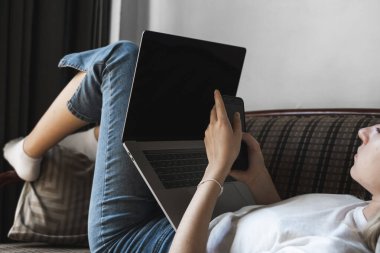 Dizüstü bilgisayarı olan bir kadın kanepede uzanıyor ve akıllı telefon kullanıyor. İnternette çalış ve çalış, serbest çalış. Defteriyle çalışan serbest çalışan bir kız elinde telefonuyla kanepede yatıyor..