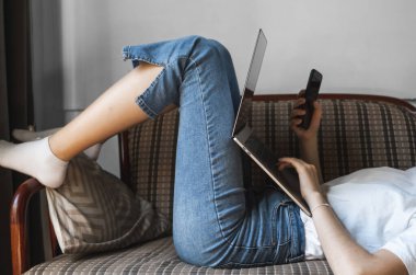 Dizüstü bilgisayarı olan bir kadın kanepede uzanıyor ve akıllı telefon kullanıyor. İnternette çalış ve çalış, serbest çalış. Defteriyle çalışan serbest çalışan bir kız elinde telefonuyla kanepede yatıyor..