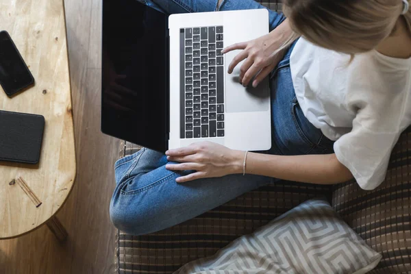 Mujer con una laptop sentada en un sofá. Estudiar y trabajar en línea, freelance. Mujer autónoma, niña que trabaja con su cuaderno sentado en un sofá con un teléfono, smartphone y lector de libros en la mesa . — Foto de Stock