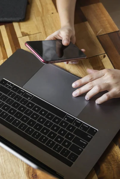 Las manos de las mujeres utilizan un teléfono inteligente con un ordenador portátil. Estudiar y trabajar en línea, freelance. Mujer por cuenta propia o independiente, chica que trabaja con su portátil sentado en la mesa de madera con un teléfono y un lector . — Foto de Stock