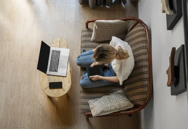Γυναίκα κάθεται σε έναν καναπέ και χρησιμοποιώντας ένα e-reader ενώ ξεκουράζεται από την εργασία με ένα φορητό υπολογιστή. Μελέτη και εργασία σε απευθείας σύνδεση, ελεύθερος επαγγελματίας. Το κορίτσι δουλεύει με το σημειωματάριό της στον καναπέ.. — Φωτογραφία Αρχείου