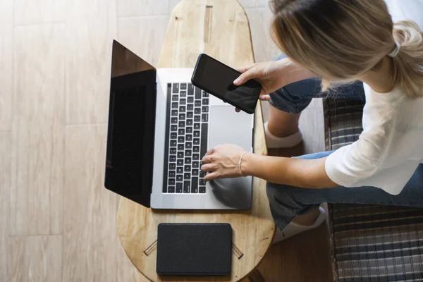 Mujer con un ordenador portátil está sentado en un sofá y utilizar un teléfono inteligente. Estudiar y trabajar en línea, freelance. Chica autónoma está trabajando con su cuaderno sentado en un sofá con un teléfono y leer en la mesa . — Foto de Stock