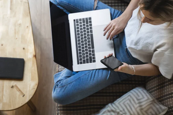 Mujer con un ordenador portátil está sentado en un sofá y utilizar un teléfono inteligente. Estudiar y trabajar en línea, freelance. Chica autónoma está trabajando con su cuaderno sentado en un sofá con un teléfono y leer en la mesa . — Foto de Stock