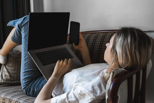 Woman with a laptop is laying on a sofa and using a smartphone. Study and work online, freelance. Self employed girl working with her notebook laying on a couch with a phone.