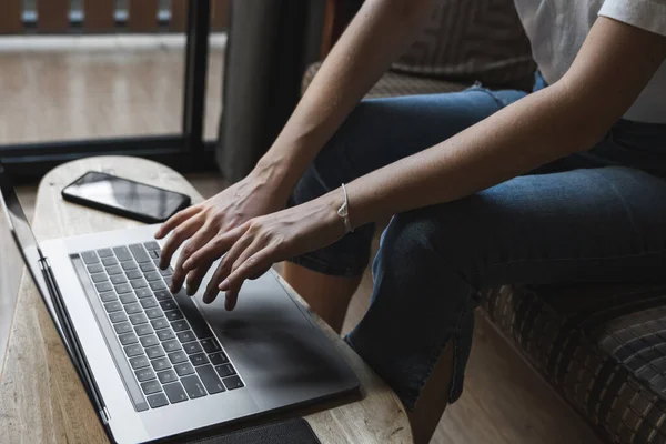 Mujer está sentado en un sofá y el uso de un ordenador portátil en la mesa de madera. Estudiar y trabajar en línea, freelance. Chica autónoma está trabajando con su cuaderno sentado en un sofá con un teléfono y leer en la mesa . — Foto de Stock