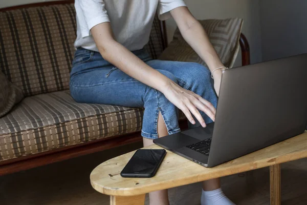 Mujer está sentado en un sofá y el uso de un ordenador portátil en la mesa de madera. Estudiar y trabajar en línea, freelance. Chica autónoma está trabajando con su cuaderno sentado en un sofá con un teléfono y leer en la mesa . — Foto de Stock