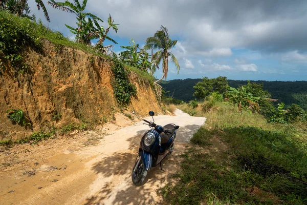 Kék robogó a földúton egy dombon a trópusi dzsungelben, festői kilátással a hegyekre egy napsütéses napon. Kaland- és utazási koncepció. — Stock Fotó